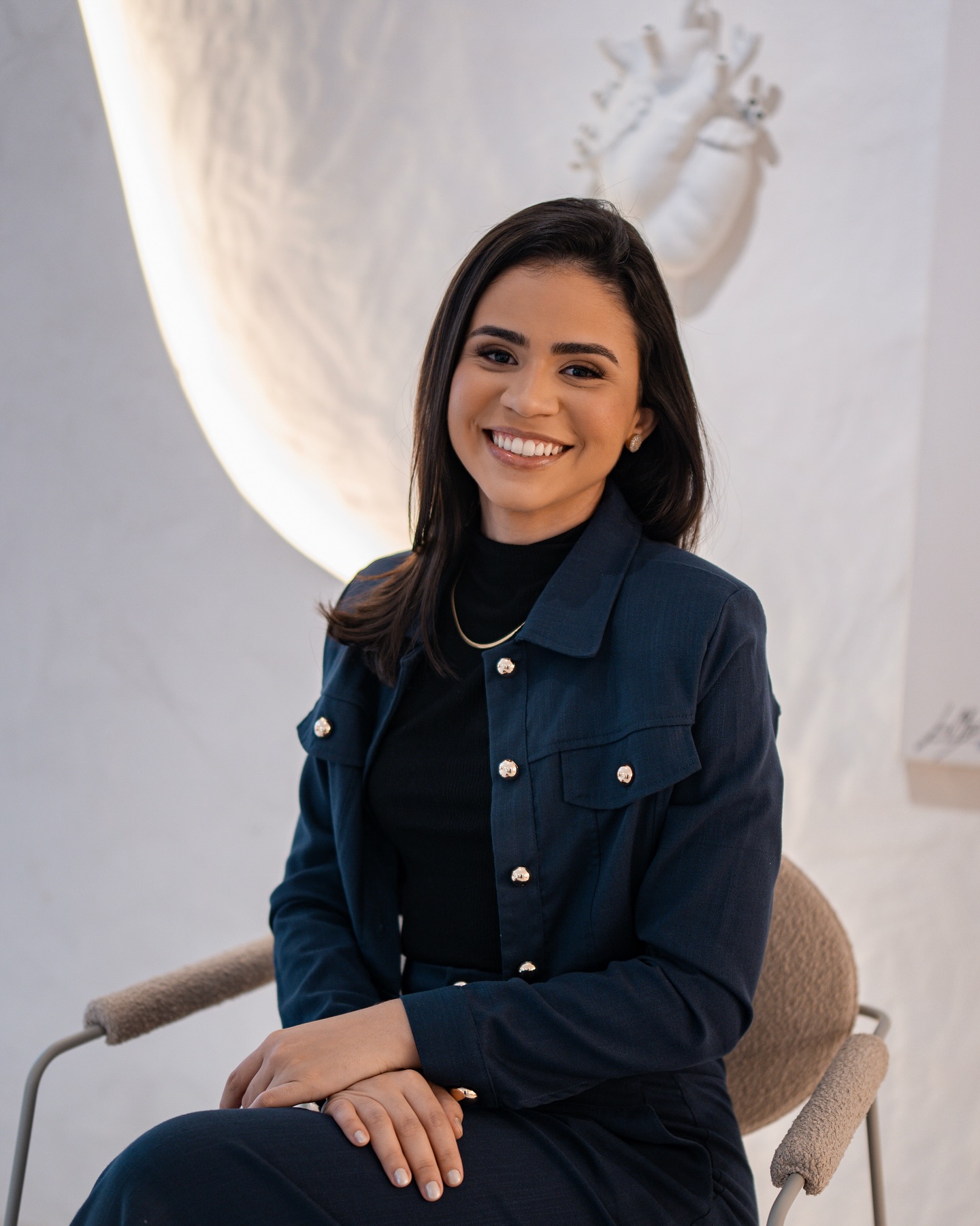 Glycia Souza sorrindo, sentada em uma poltrona azul ao lado de uma planta